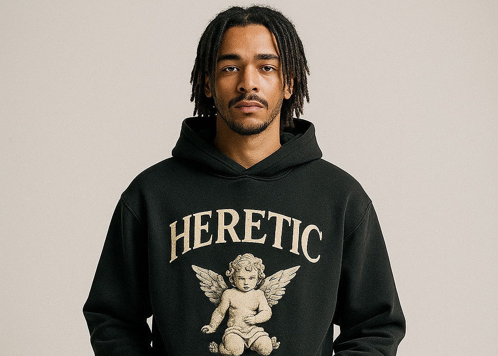 Person wearing a black hoodie with 'HERETIC' and an angel graphic on a beige background
