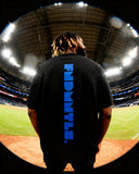 Model in black Mets Blue T-Shirt - Heavyweight Earth Tone showing bold vertical FNDMTLS logo at stadium. Unisex cotton tee.