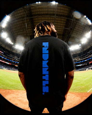 Model in black Mets Blue T-Shirt - Heavyweight Earth Tone showing bold vertical FNDMTLS logo at stadium. Unisex cotton tee.
