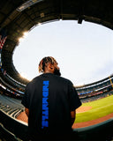 Back view of black Mets Blue T-Shirt - Heavyweight Earth Tone with vertical blue FNDMTLS logo. Styled for stadium streetwear.