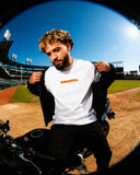 White Mets Orange Heavyweight T-Shirt with orange FNDMTLS logo, styled on a motorcycle at a baseball field for streetwear vibes.