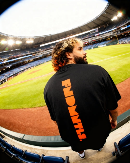 Black Mets Orange Heavyweight T-Shirt with vertical orange FNDMTLS logo on back, worn at stadium for casual streetwear appeal.