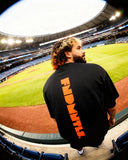 Black Mets Orange Heavyweight T-Shirt with vertical orange FNDMTLS logo on back, worn at stadium for casual streetwear appeal.