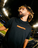 Mets Orange Heavyweight T-Shirt in black with bold orange FNDMTLS logo, worn at a baseball stadium. Unisex cotton streetwear top.
