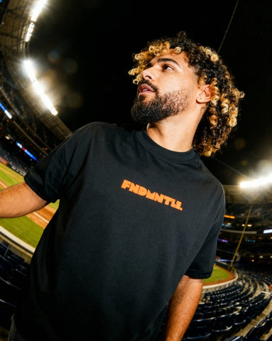 Mets Orange Heavyweight T-Shirt in black with bold orange FNDMTLS logo, worn at a baseball stadium. Unisex cotton streetwear top.