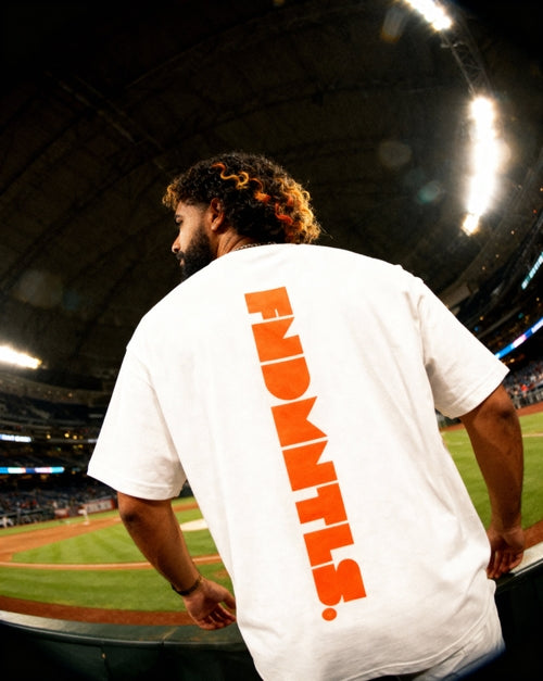 Close-up of white Mets Orange Heavyweight T-Shirt with vertical orange FNDMTLS logo, worn at baseball stadium for casual fashion.