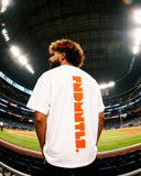 Vertical orange FNDMTLS logo on back of white Mets Orange Heavyweight T-Shirt, worn at stadium for casual MLB streetwear style.