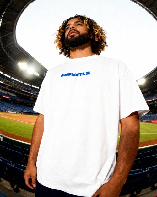 Mets Blue T-Shirt - Heavyweight Earth Tone in white with bold FNDMTLS logo, worn at a baseball stadium. Unisex cotton casual streetwear.