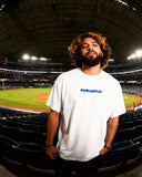 Model in Mets Blue T-Shirt - Heavyweight Earth Tone in white, standing in stadium seating. Features bold FNDMTLS logo on front.